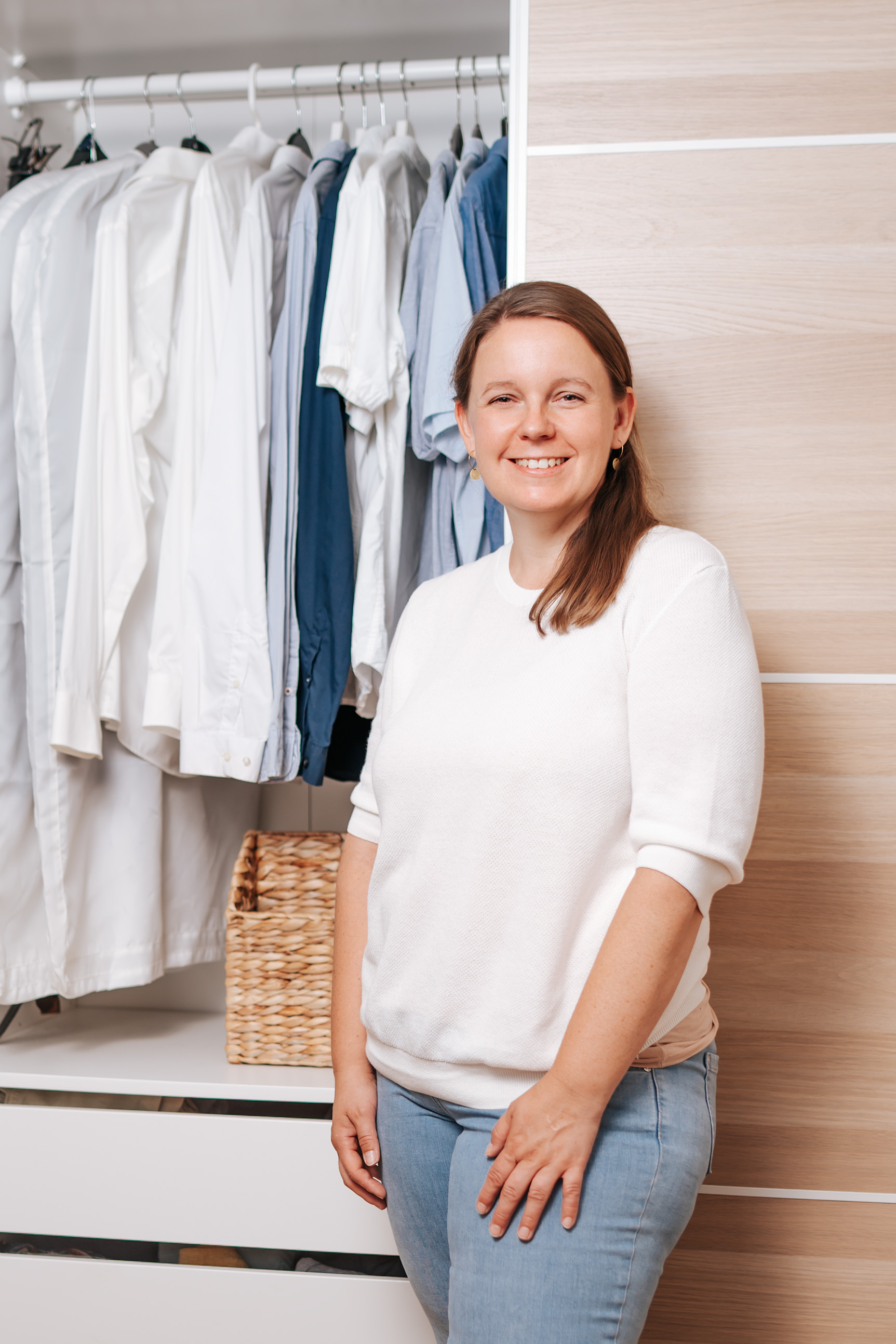 Die Frau steht vor einem Schrank, in dem noch alles ordentlich aufgehängt ist. Sie trägt Jeans und ein weißes Pullover. De...